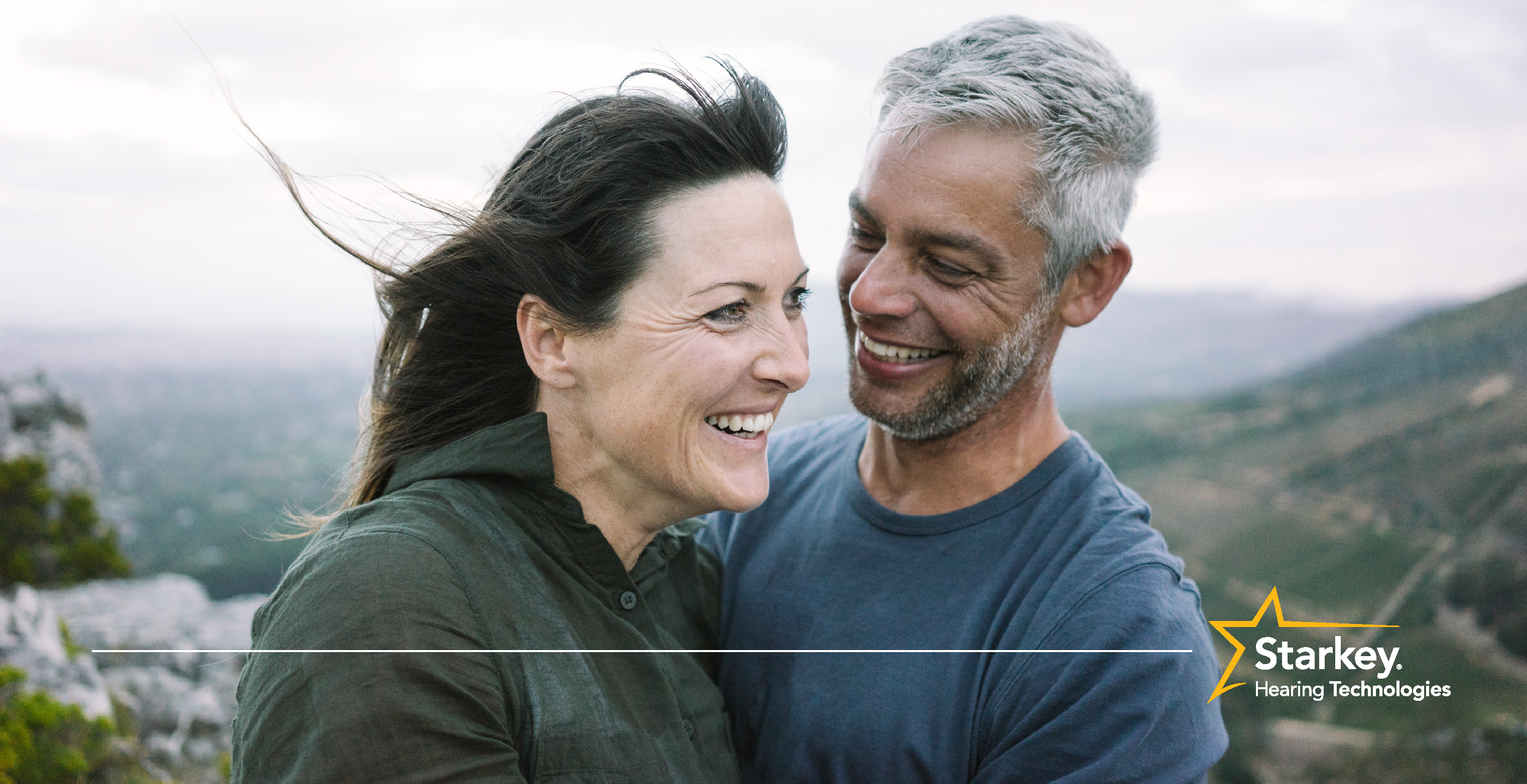 Homme aux cheveux gris souriant debout à côté d'une femme aux cheveux bruns souriant sur une falaise